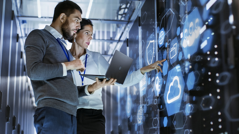 male it specialist holds laptop and discusses work with female server technician. they're standing in data center, rack server cabinet with cloud server icon and visualization.